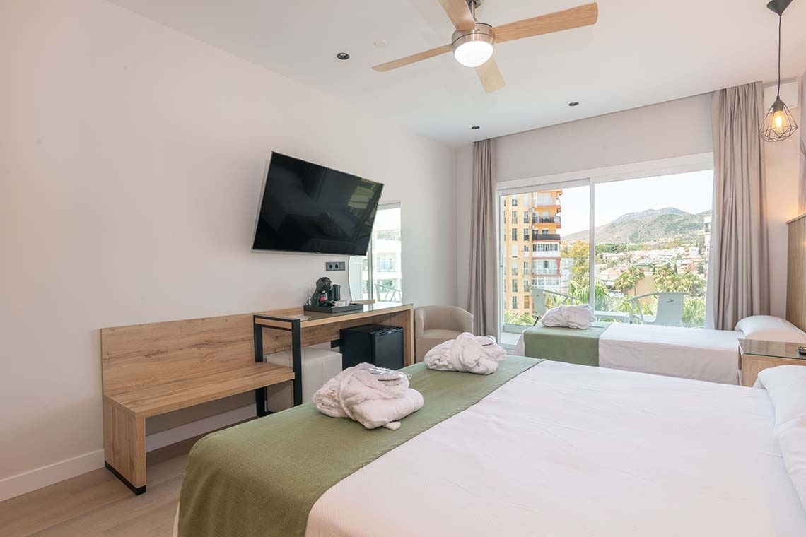 Una moderna habitación de hotel con dos camas, un televisor, escritorio con cafetera y minibar, y un balcón con vistas a la ciudad y las montañas.