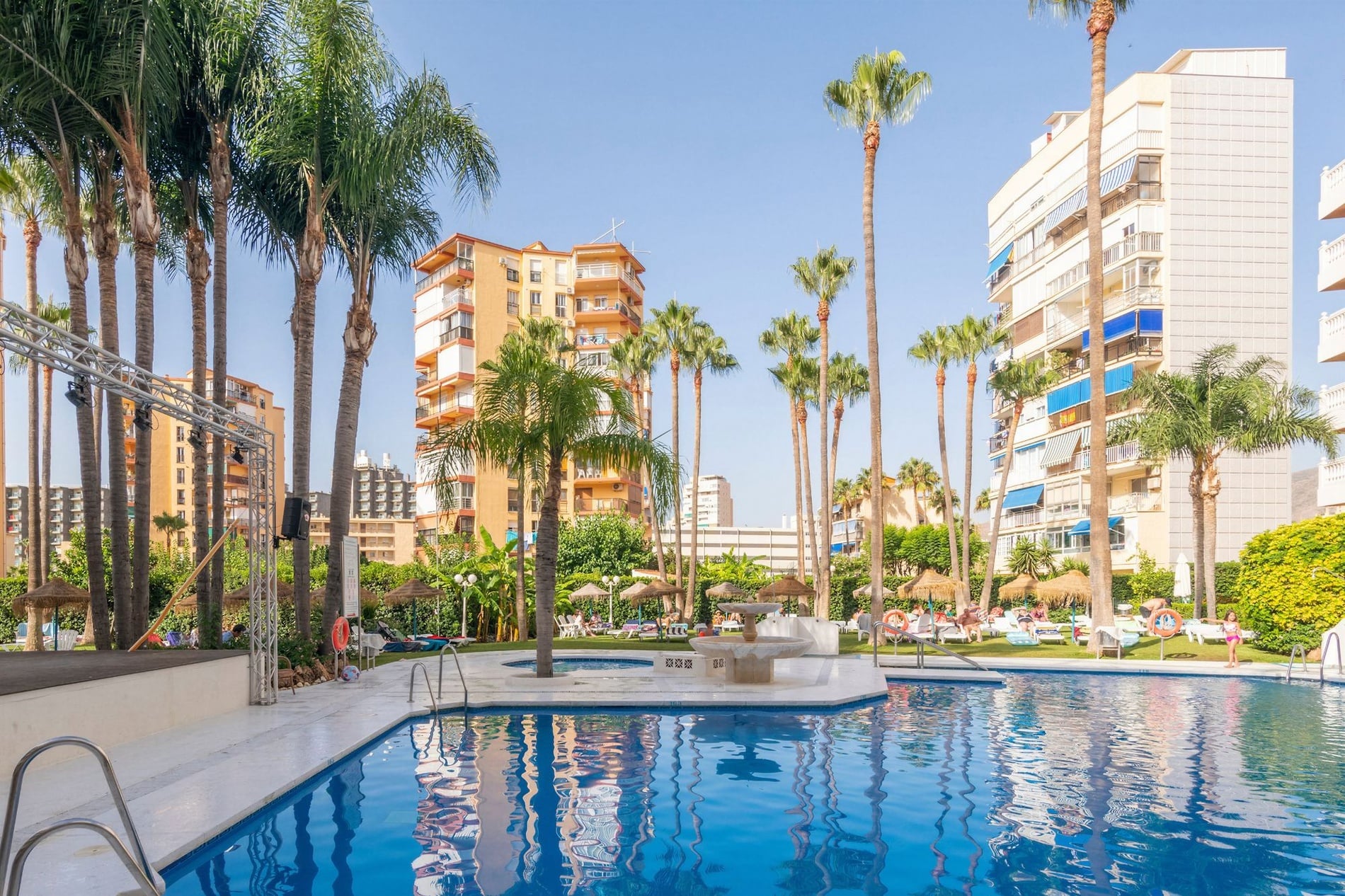 Una vista de un área de piscina al aire libre rodeada de palmeras, edificios residenciales y tumbonas bajo un cielo azul claro.