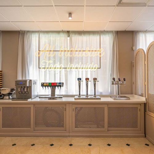 Mostrador de bebidas de autoservicio con dispensadores variados y un luminoso de neón 'All Inclusive / Todo Incluido' sobre un fondo de cortinas claras.