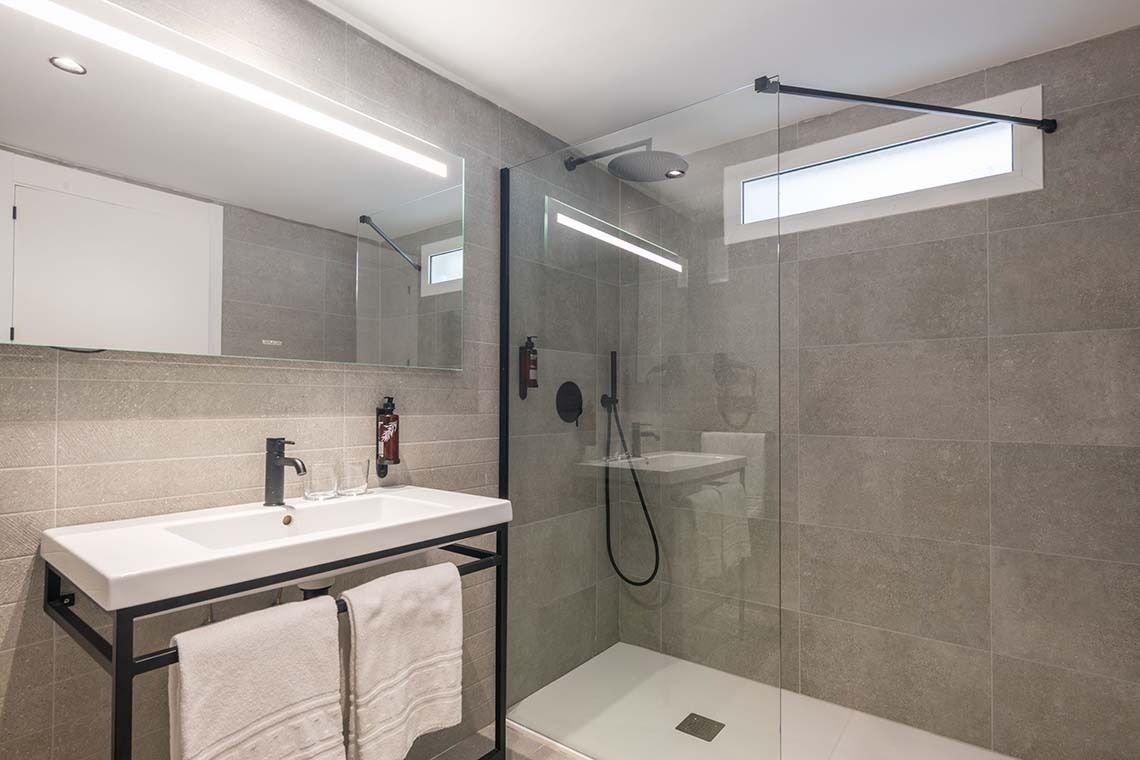 Baño moderno con paredes de azulejos grises, un lavabo blanco con grifo negro, un espejo grande con iluminación LED y una ducha a ras de suelo con mampara de cristal.