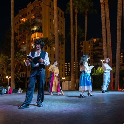 Cuatro actores ensayan o actúan en un escenario al aire libre por la noche, rodeados de palmeras y edificios iluminados en el fondo.