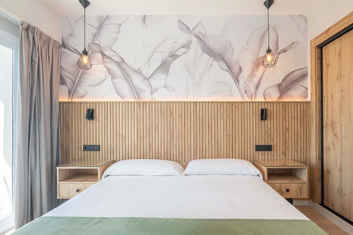 Una habitación moderna y luminosa con una cama doble, un cabecero de lamas de madera, papel pintado con grandes hojas monocromáticas y mesitas de noche flotantes.