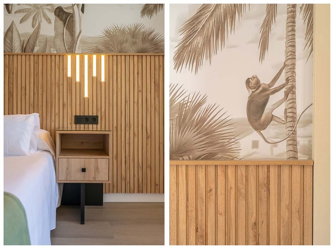 Una habitación de hotel moderna presenta una cabecera de madera laminada con iluminación LED integrada y papel tapiz tropical con palmeras y un mono escalando un árbol, junto a una mesita de noche.
