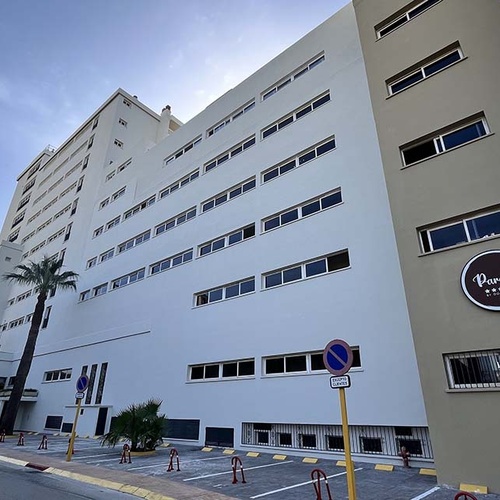 Se muestra un edificio alto de color claro con un letrero que dice 'Parasol **** by Dopore', flanqueado por palmeras y una calle con marcas de aparcamiento, bajo un cielo azul.
