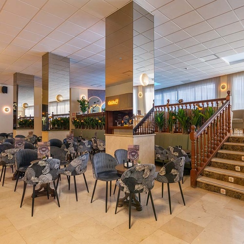 Una sala de hotel o vestíbulo con un área de asientos de estilo moderno, columnas espejadas, una elegante escalera de madera y grandes ventanales con cortinas traslúcidas.