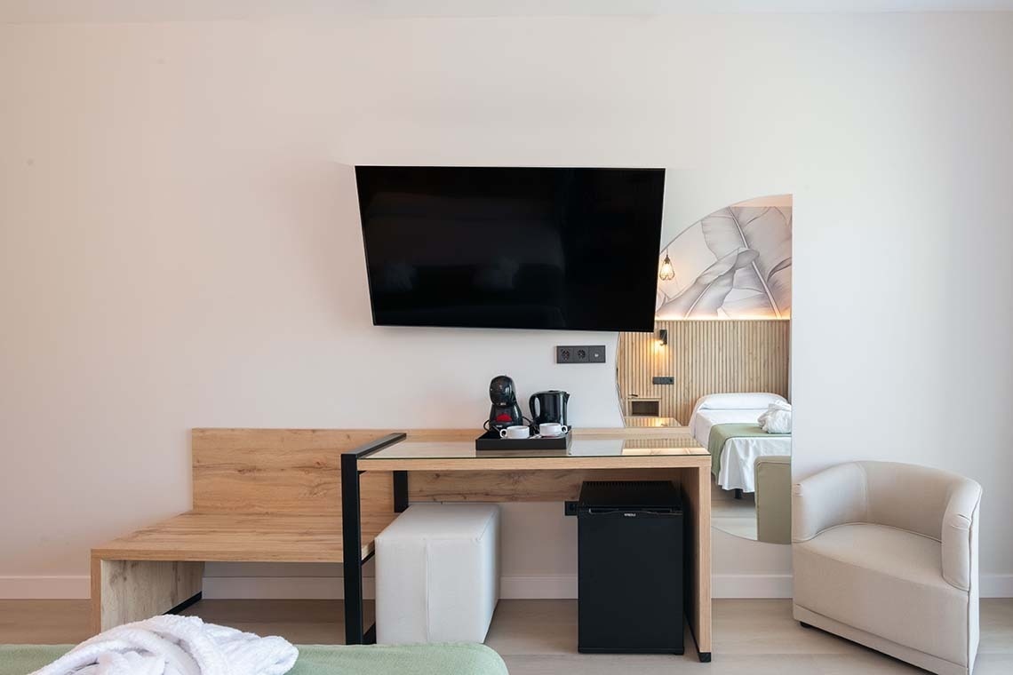 En una habitación de hotel moderna, una televisión cuelga sobre un escritorio con cafetera y minibar, mientras una cama y un sillón se vislumbran en una alcoba adyacente.
