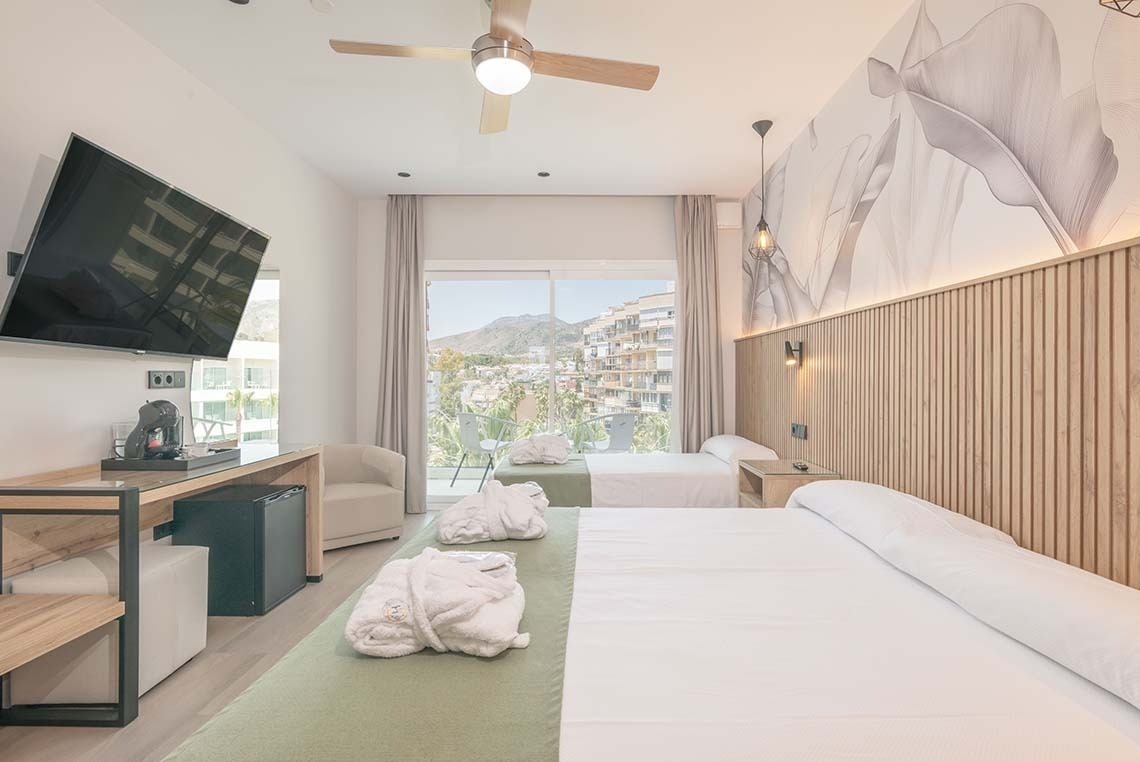 Una moderna habitación de hotel cuenta con dos camas, un televisor montado en la pared, un ventilador de techo y un balcón con vistas a edificios y montañas.