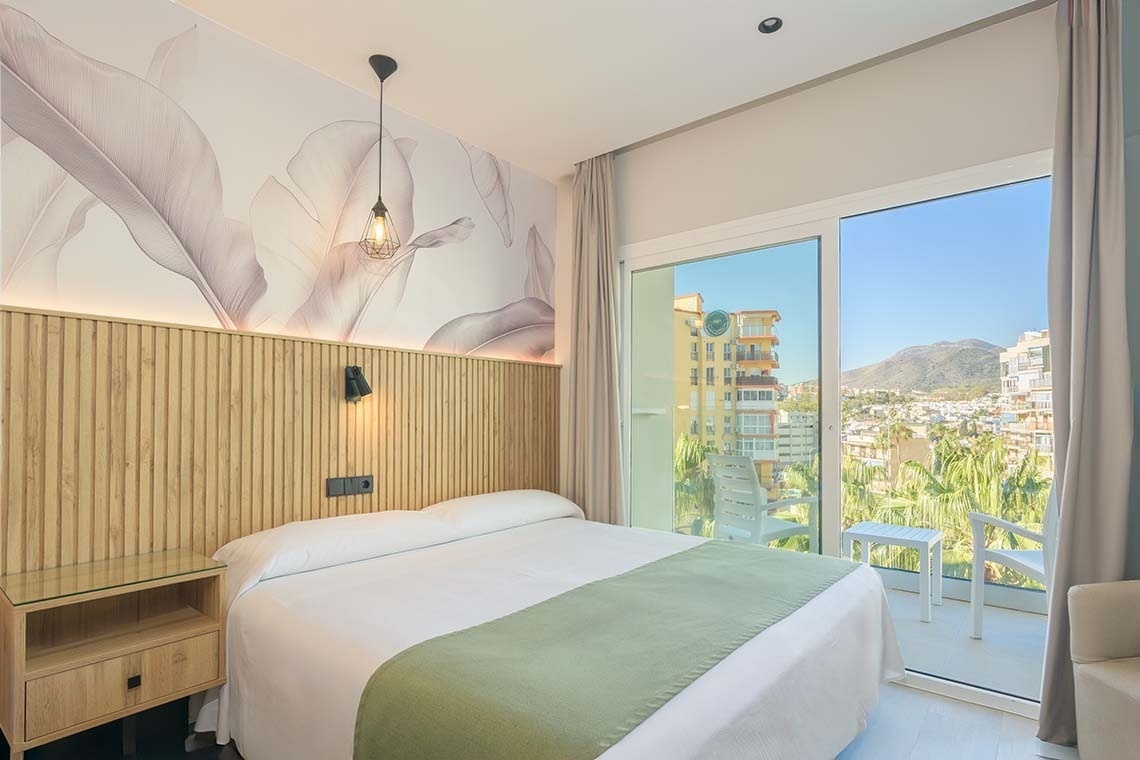 Habitación de hotel moderna y luminosa con cama, cabecero de madera y papel pintado botánico, y balcón con vistas a la ciudad y montañas.
