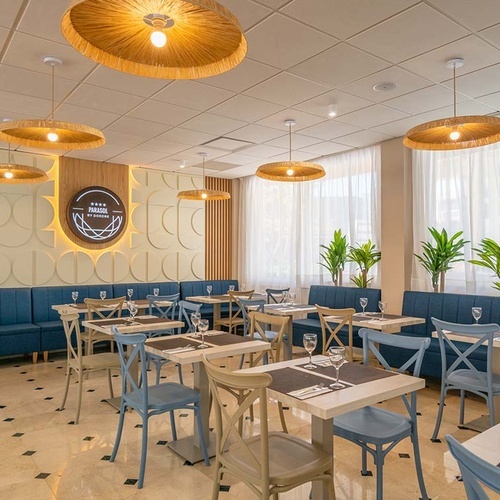 Un restaurante moderno y luminoso presenta mesas con sillas de varios colores, asientos tipo banco tapizados en azul, una pared decorada con un patrón geométrico y el logo 