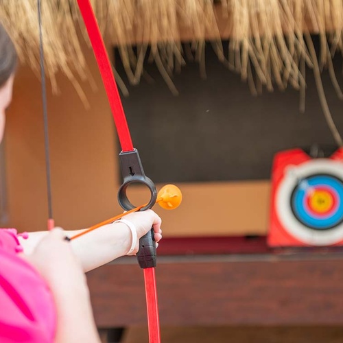 Una persona apunta con un arco de juguete rojo una flecha naranja con punta de bola a una diana.