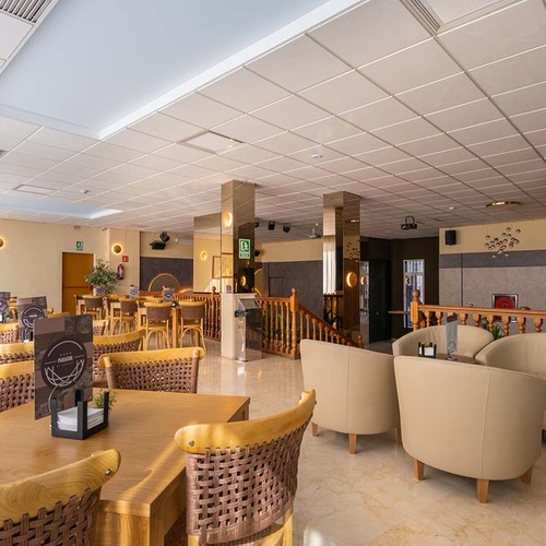 Un amplio y luminoso salón interior con mesas de madera, sillas de mimbre, sillones tapizados, pilares espejados y una barandilla de madera.