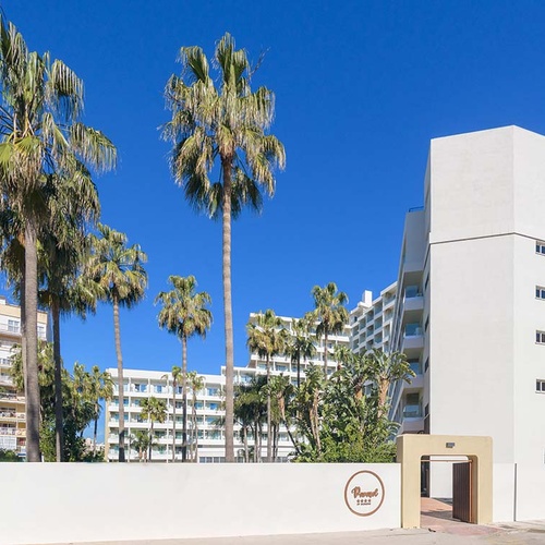 Un edificio blanco moderno con múltiples palmeras se alza bajo un cielo azul brillante, con una entrada distintiva que lleva el nombre 