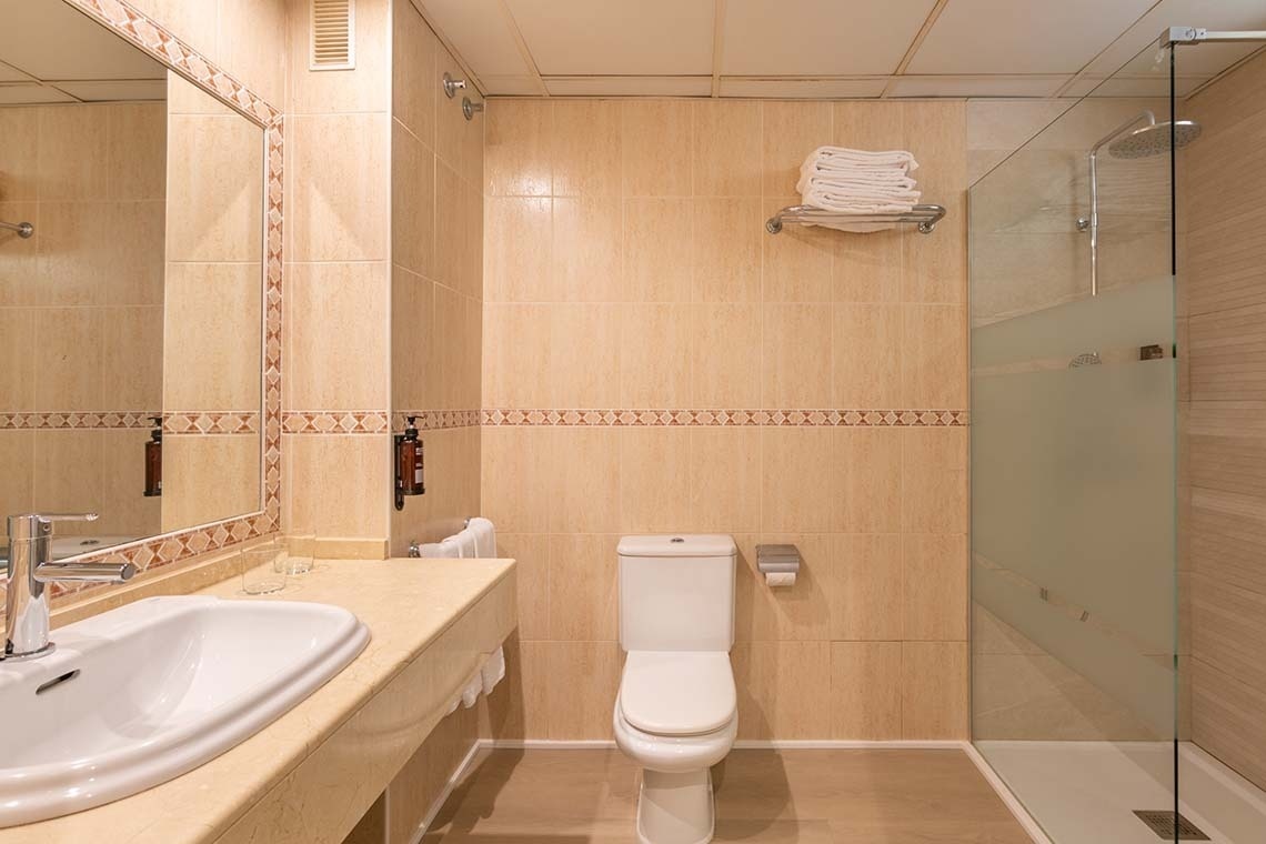 Este es un cuarto de baño con azulejos beige, un lavabo con espejo, un inodoro y una ducha con mampara de cristal.