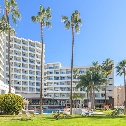 Un complejo hotelero moderno con edificios blancos, palmeras, un césped verde y una piscina, bajo un cielo azul despejado.