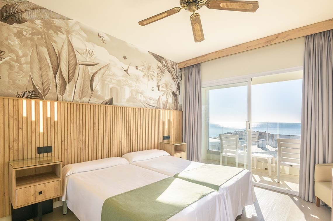 Habitación de hotel luminosa con dos camas, un mural tropical en la pared, un ventilador de techo y un balcón con vistas al mar.