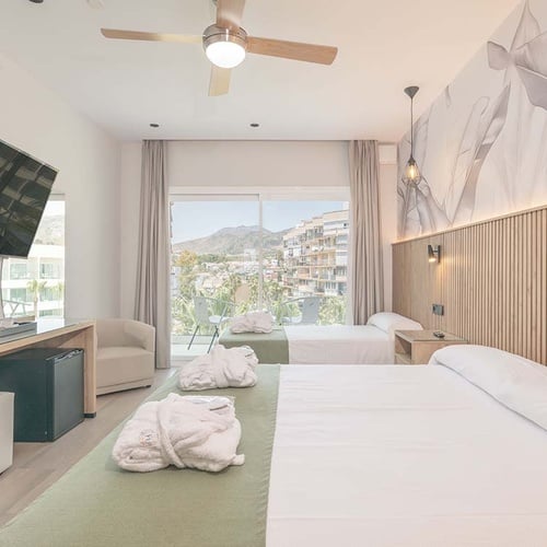 Habitación de hotel moderna y bien equipada con dos camas, televisor, cafetera, minibar y un balcón con vistas a la ciudad y las montañas.