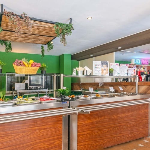 Una estación de autoservicio con un buffet de ensaladas y una decoración tropical de bambú, con un bar al fondo.