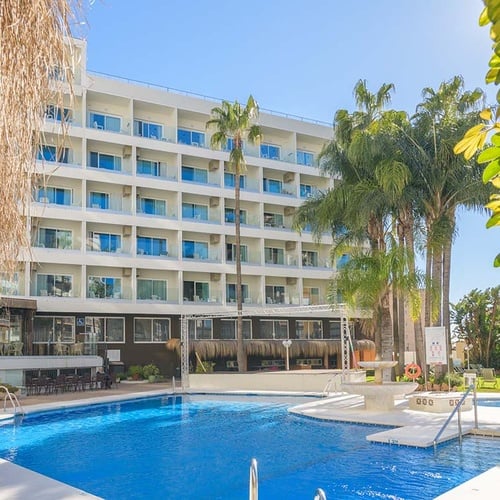 La imagen muestra un hotel de varias plantas con balcones y palmeras, junto a una piscina azul brillante bajo un cielo soleado.
