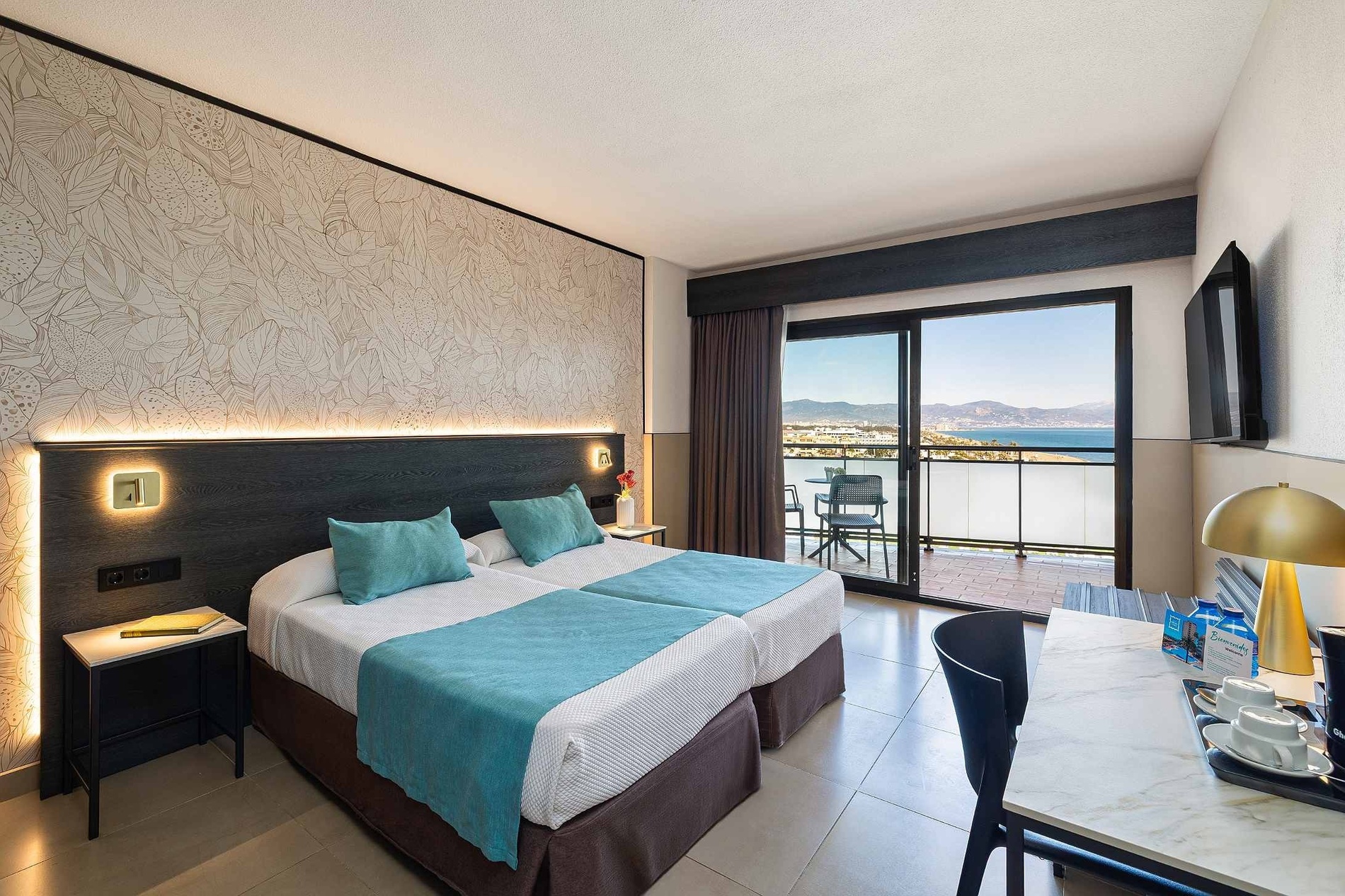 Une chambre d'hôtel lumineuse et moderne avec deux lits, un balcon offrant une vue imprenable sur la mer et les montagnes, et un coin bureau.