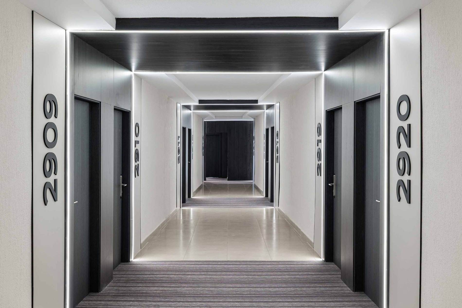 Un couloir d'hôtel moderne et symétrique est illuminé par des lignes lumineuses à LED verticales et horizontales, encadrant des portes sombres et affichant des numéros de chambre en relief sur des murs texturés.
