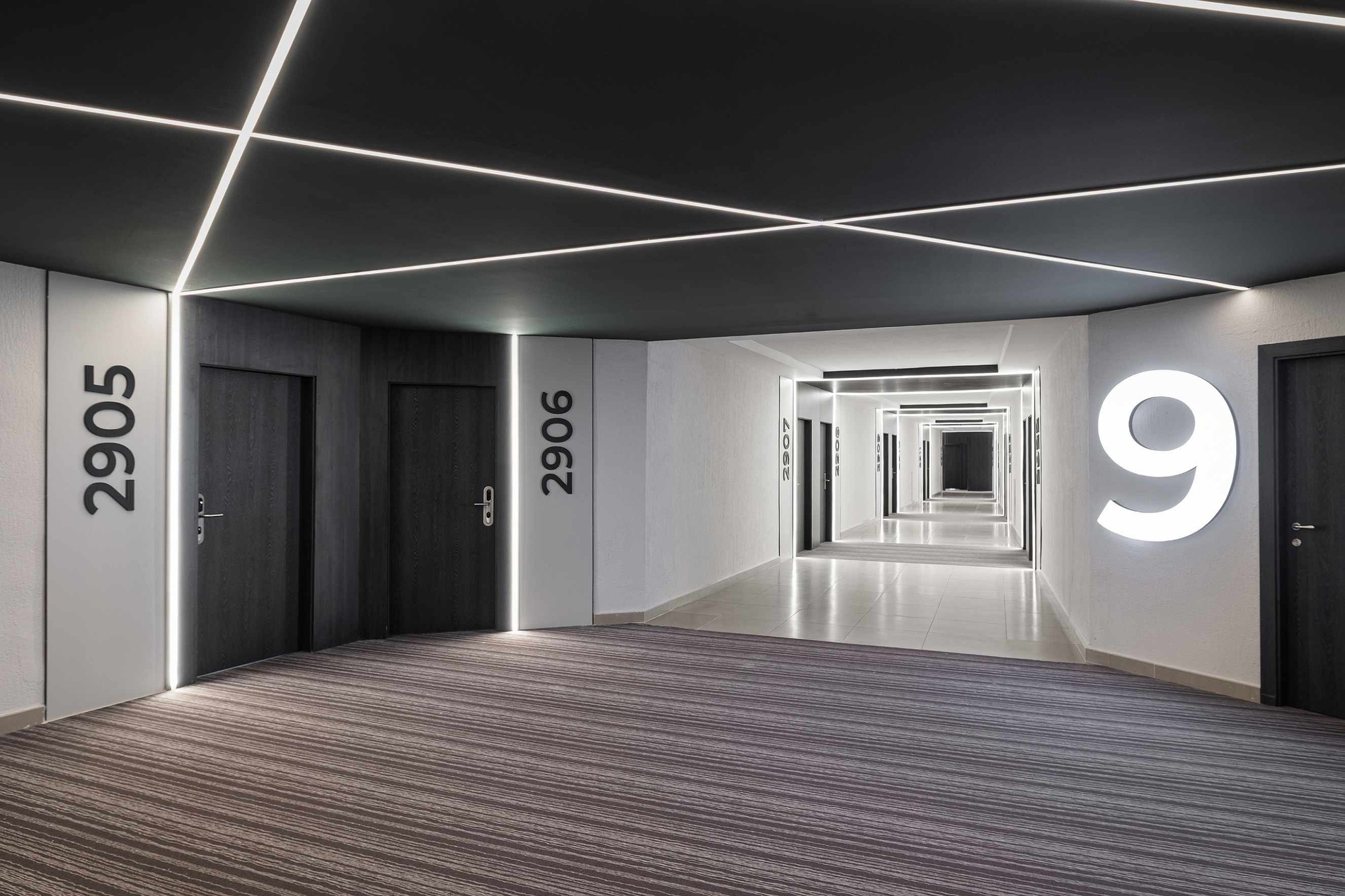 Un couloir d'hôtel moderne présente un plafond noir orné de bandes lumineuses LED blanches entrecroisées, des portes sombres avec des numéros de chambre comme 