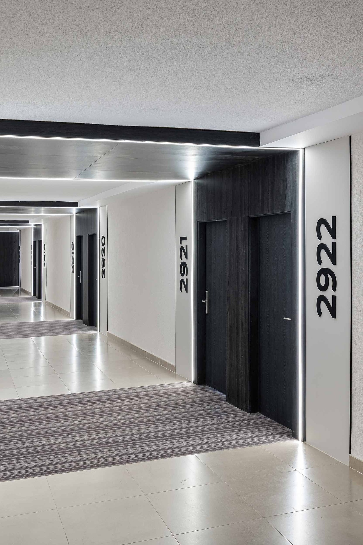 Un couloir d'hôtel au design moderne présente des portes sombres numérotées, éclairées par des bandes lumineuses verticales et un éclairage linéaire au plafond, le tout sur un sol carrelé et un tapis à rayures.