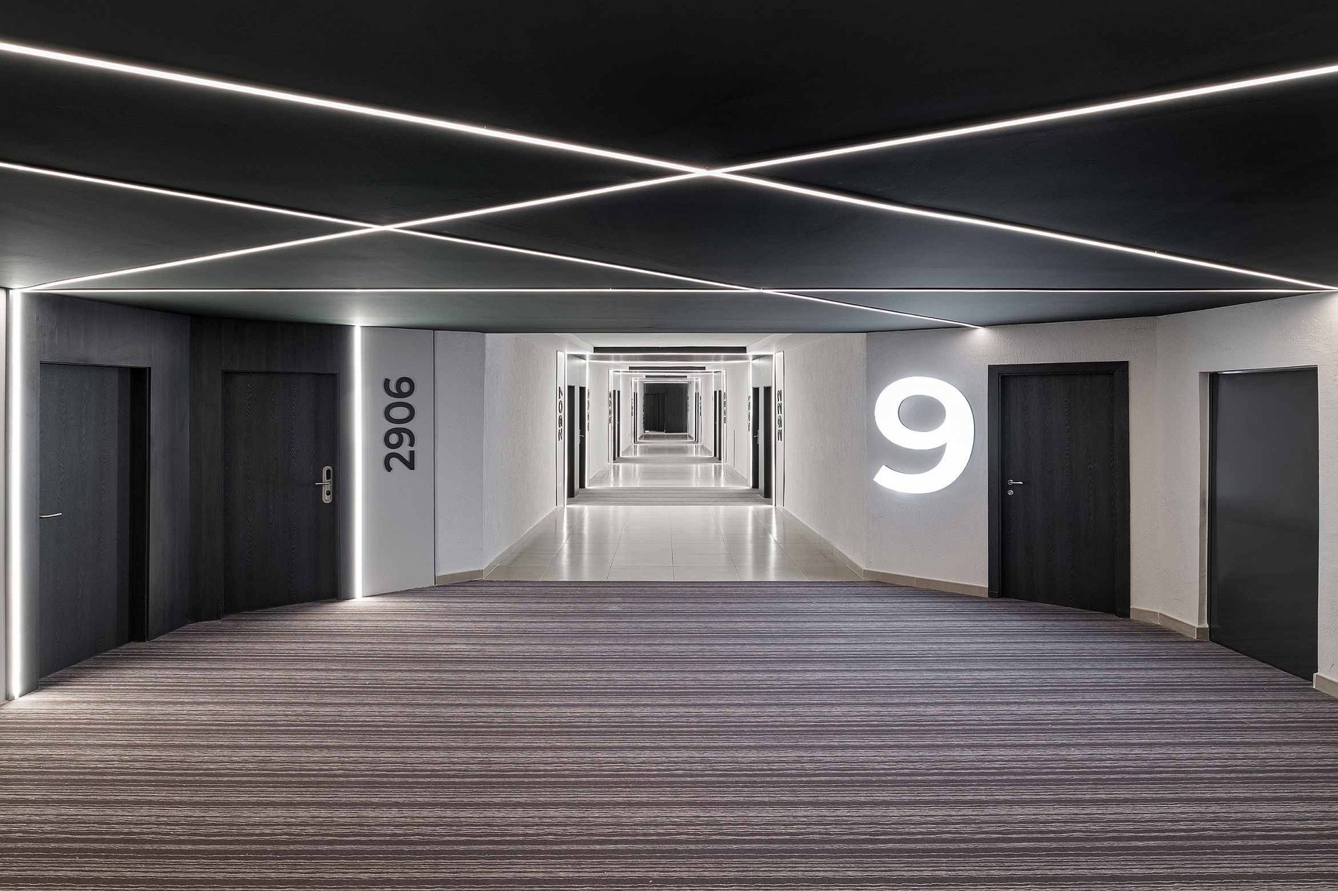 Un couloir d'hôtel moderne et minimaliste présente un plafond noir avec des bandes lumineuses LED croisées et un tapis à rayures qui s'étend en perspective.