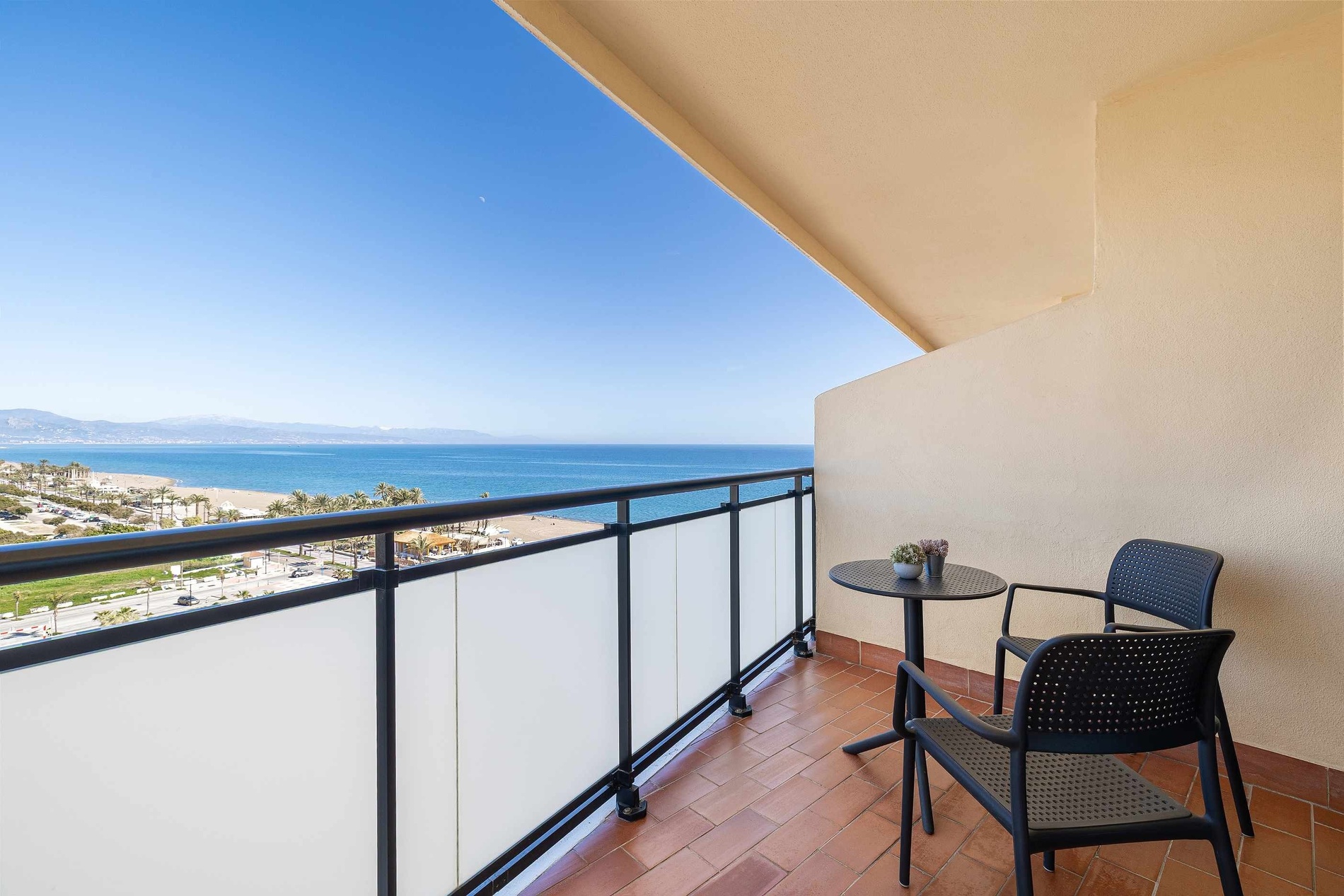 Un balcon ensoleillé offre une vue imprenable sur la mer Méditerranée, la plage et les montagnes au loin, agrémenté d'une table et de chaises.