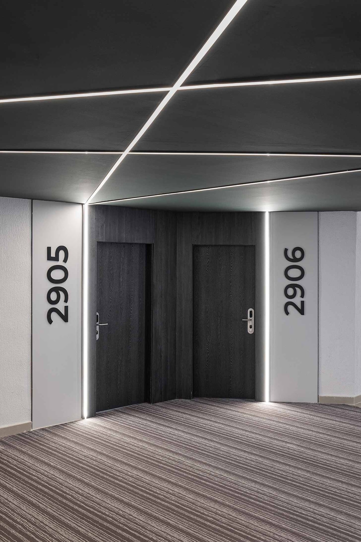Un couloir d'hôtel moderne présente deux portes sombres numérotées 2905 et 2906, éclairées par des bandes LED linéaires au plafond et verticalement.