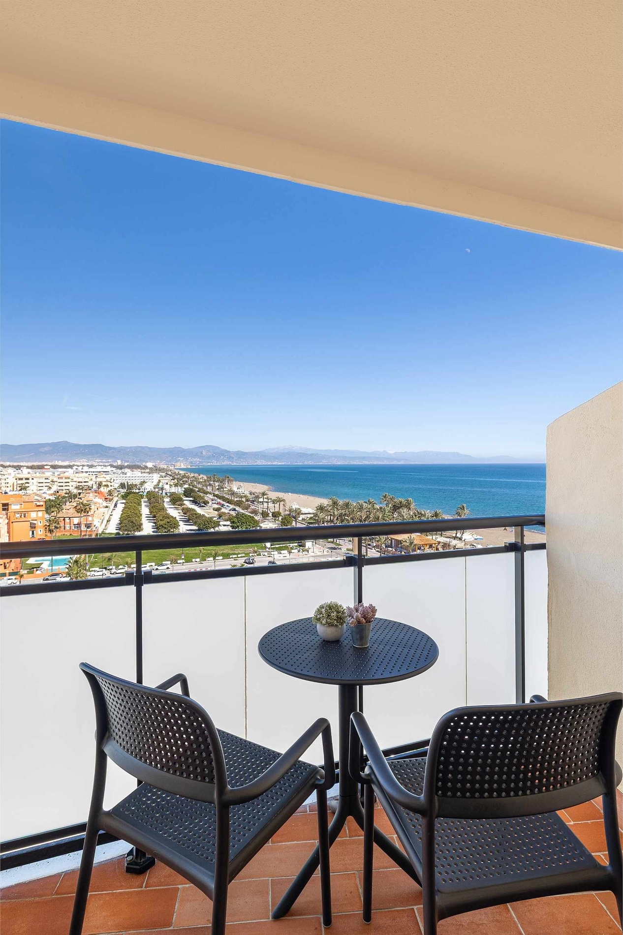 fr: Depuis un balcon meublé d'une table et deux chaises, l'image offre une vue dégagée sur une plage, la mer, une ville côtière et des montagnes lointaines sous un ciel bleu clair.