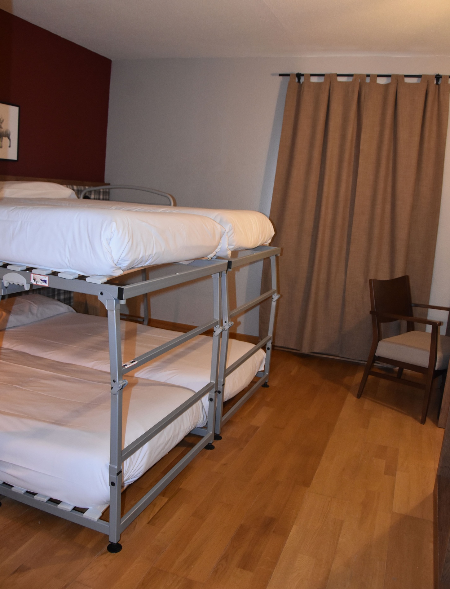 A room features a modern metal-framed bunk bed with white bedding, set against a dark red accent wall and light grey walls, with hardwood floors, brown curtains, and a wooden chair visible.