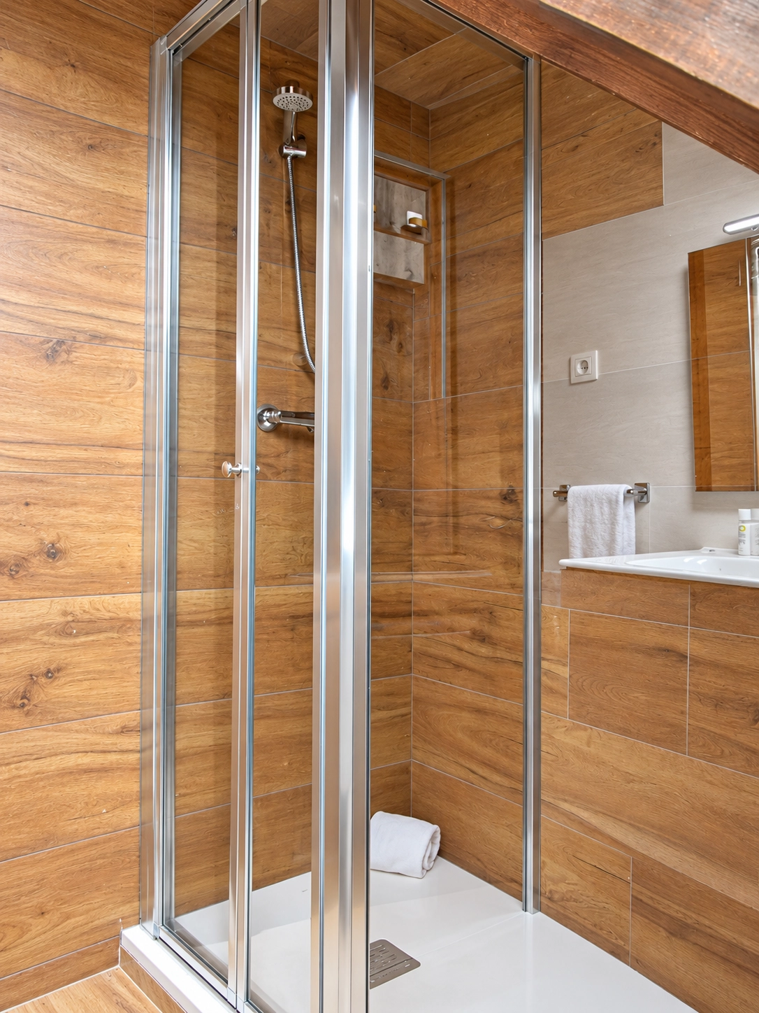 Ducha moderna con mampara de cristal, azulejos efecto madera y accesorios de lujo. SNÖ Vall de Boi en baño de hotel.