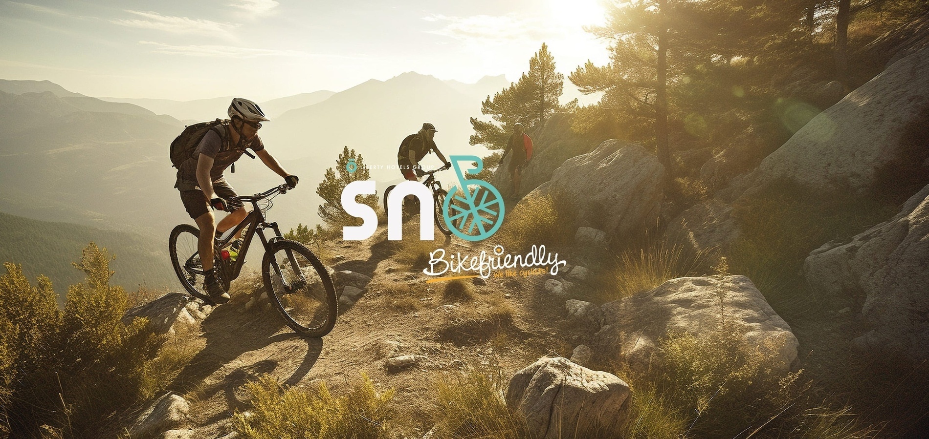 La imagen muestra a ciclistas de montaña en un sendero rocoso con montañas de fondo y una luz brillante del sol, junto a los logotipos "sn" y "Bikefriendly".