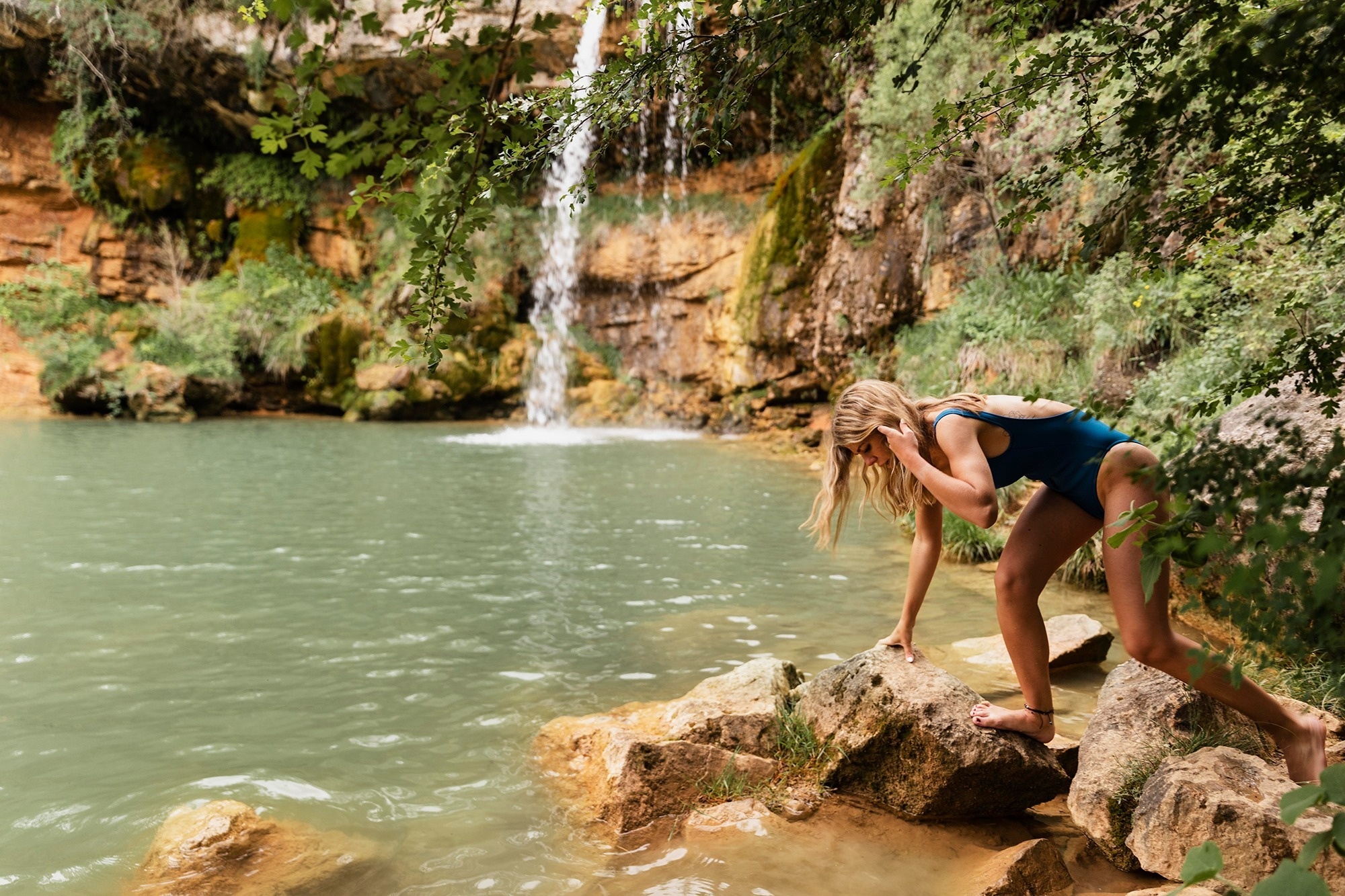 
                                    a woman in a blue swimsuit stands on a rock near a waterfall