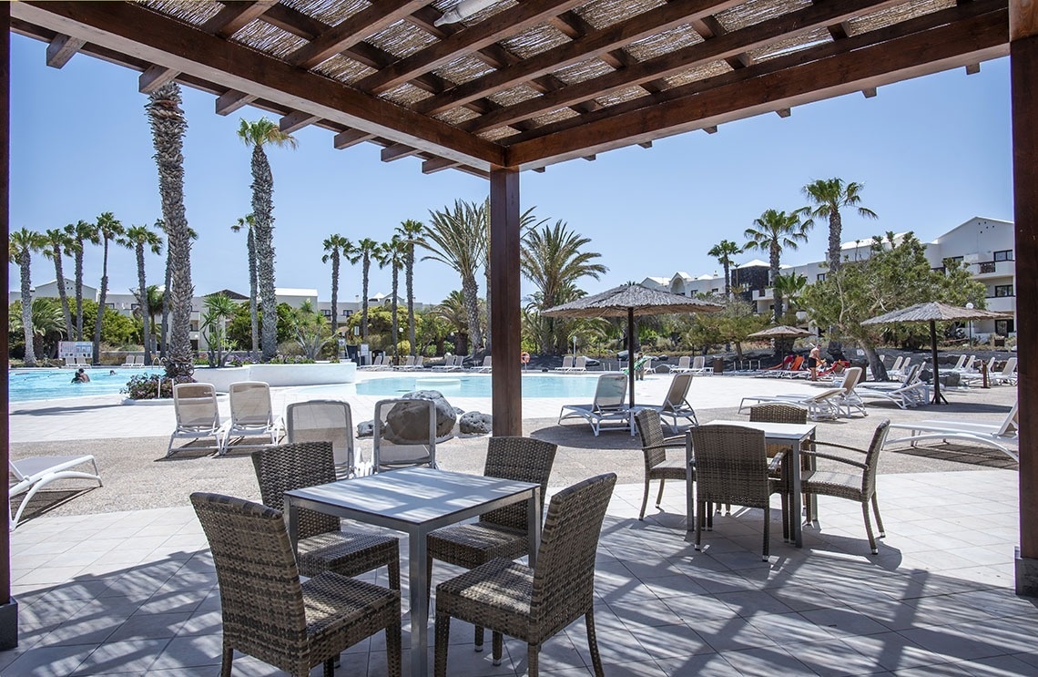 Sonniger Hotelpoolbereich mit schattiger Terrasse, Palmen und Liegestühlen.