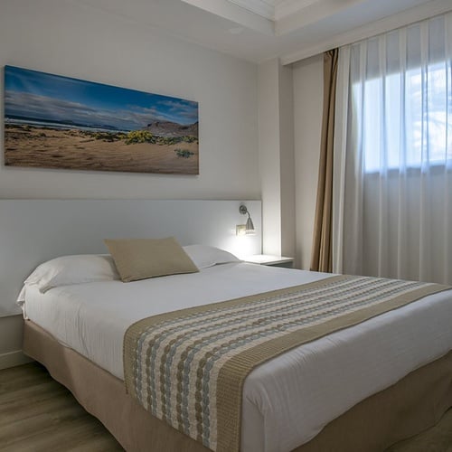 Helles, modernes Hotelzimmer im Impressive Zocos Lanzarote mit gemütlichem Bett, Strandbild und Fenster.