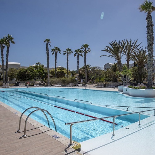 Großer Pool im Impressive Zocos Lanzarote mit Palmen, Liegestühlen und Hotelgebäuden unter blauem Himmel.