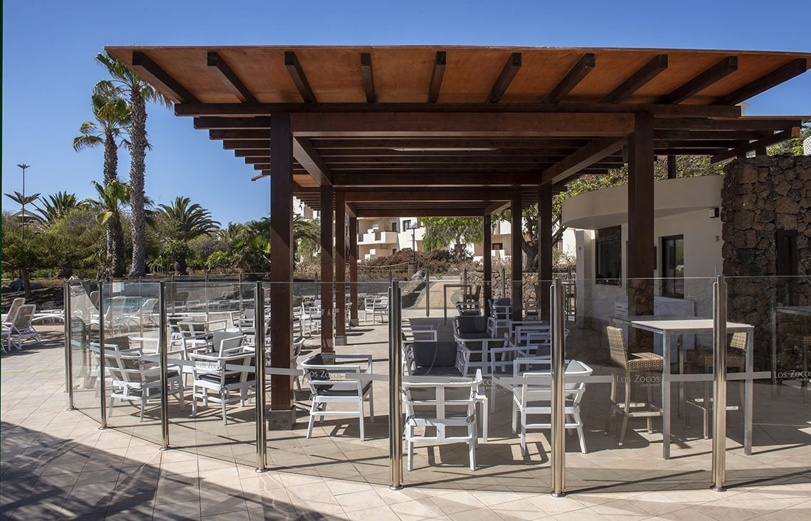 Los Zocos Resort: Sonnige Poolterrasse mit Holzpergola, gemütlichen Tischen & Stühlen unter Palmen.