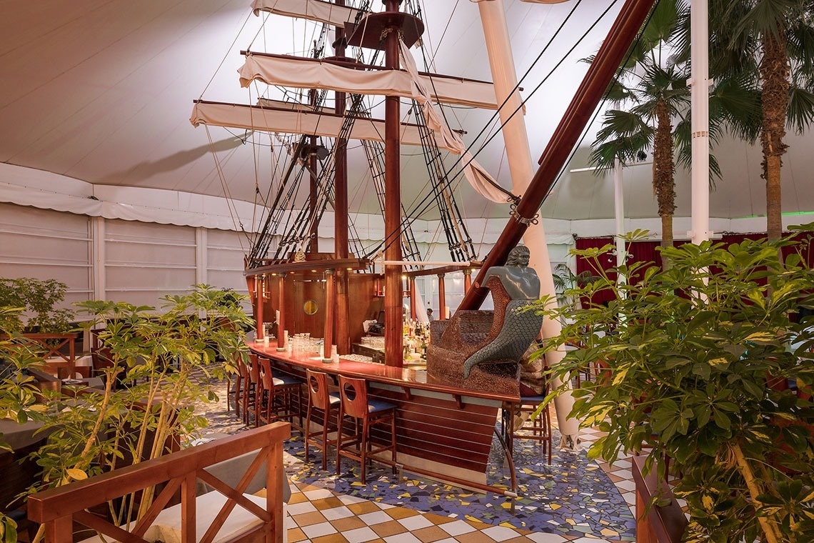 Thematische Hotelbar im Schiffsdesign mit Meerjungfrau, Palmen und tropischem Flair unter Zeltdecke.
