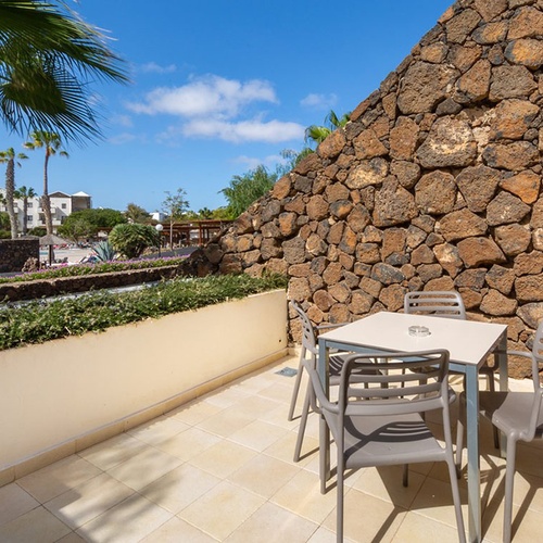Sonniger Balkon im Impressive Zocos Lanzarote mit Sitzbereich, Steinmauer und Blick auf Palmen und Hotelanlage.