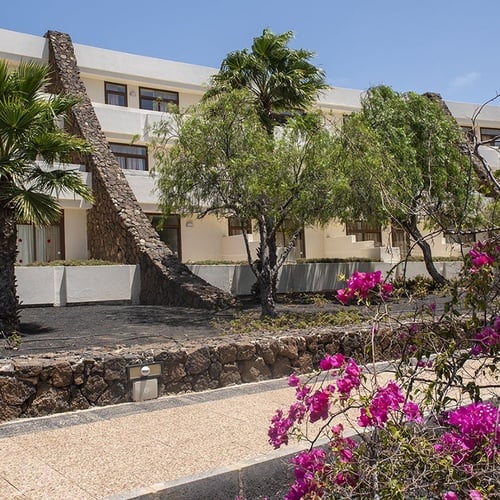 Impressive Zocos Lanzarote: Hotelgebäude mit Palmen, Steinmauer und pinker Bougainvillea unter blauem Himmel.
