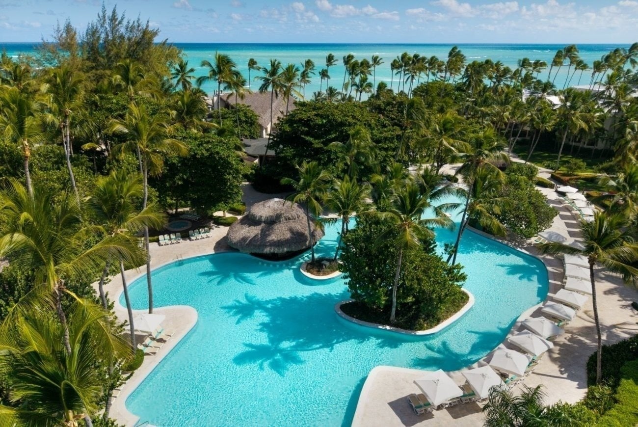 An aerial view captures a sprawling tropical resort featuring a large, irregularly shaped light blue swimming pool surrounded by abundant palm trees and lush greenery, with the turquoise ocean visible in the background.