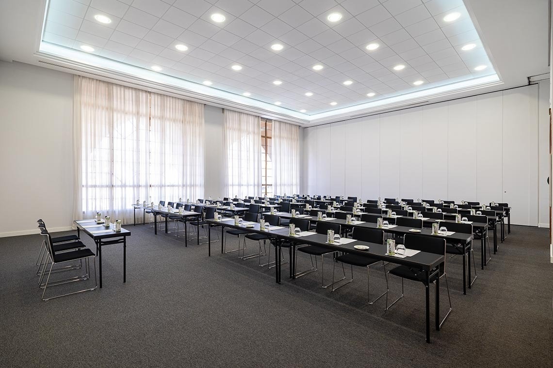 A spacious, well-lit conference room is set up with multiple rows of black tables and chairs, each with place settings, arranged on a dark carpeted floor, with large windows covered by sheer white curtains on one side and a modular white wall on the other.