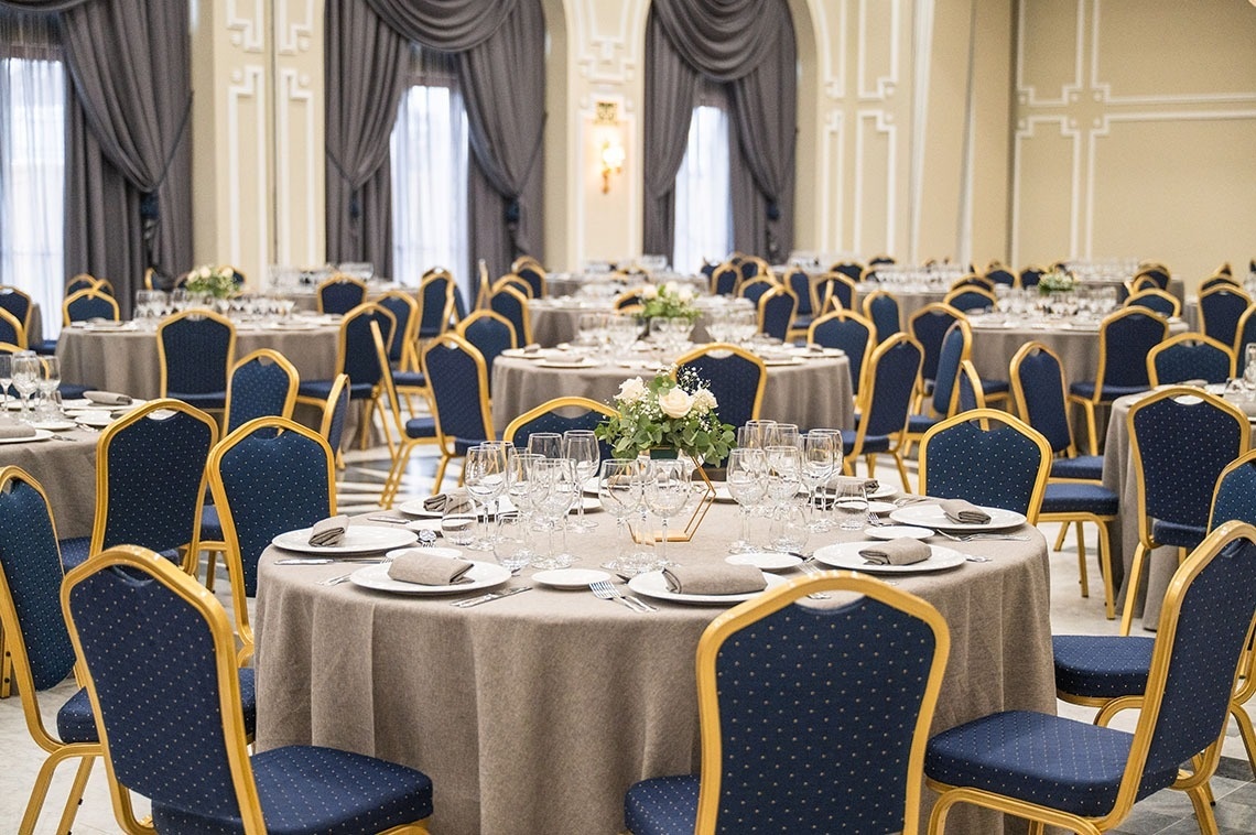 Une grande salle de banquet est élégamment dressée avec de nombreuses tables rondes recouvertes de nappes grises, entourées de chaises bleues et dorées, et décorées de centres de table floraux, prête pour un événement.