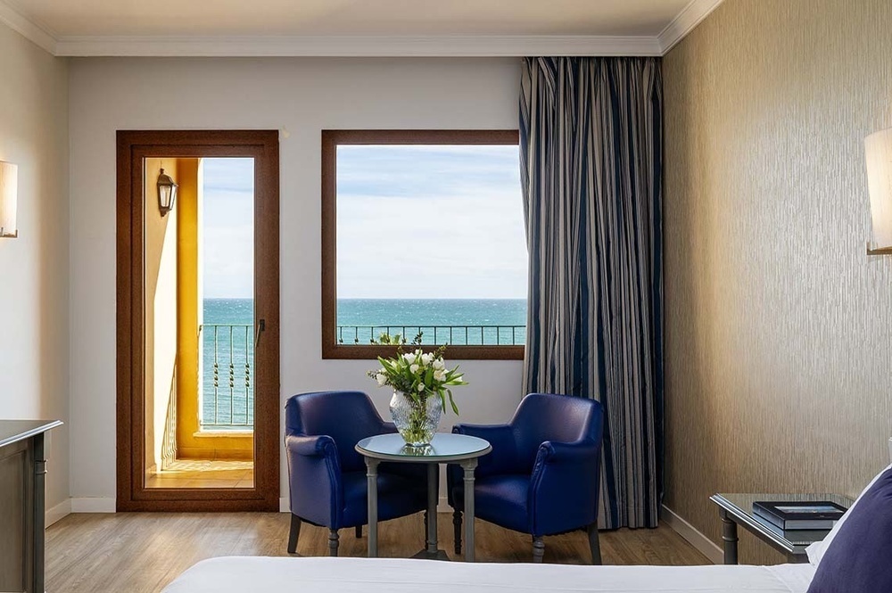 Ein helles Zimmer mit Meerblick, ausgestattet mit einer Balkontür, einem Fenster und einer Sitzecke mit zwei blauen Sesseln und einem Tisch mit Blumen.