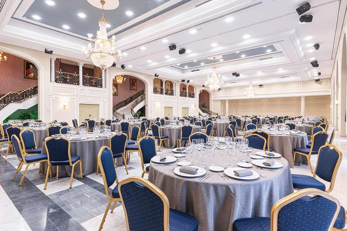 Une grande salle de réception luxueuse est aménagée pour un événement, présentant de multiples tables rondes dressées, des chaises bleues et or, sous de grands lustres et des balcons majestueux.