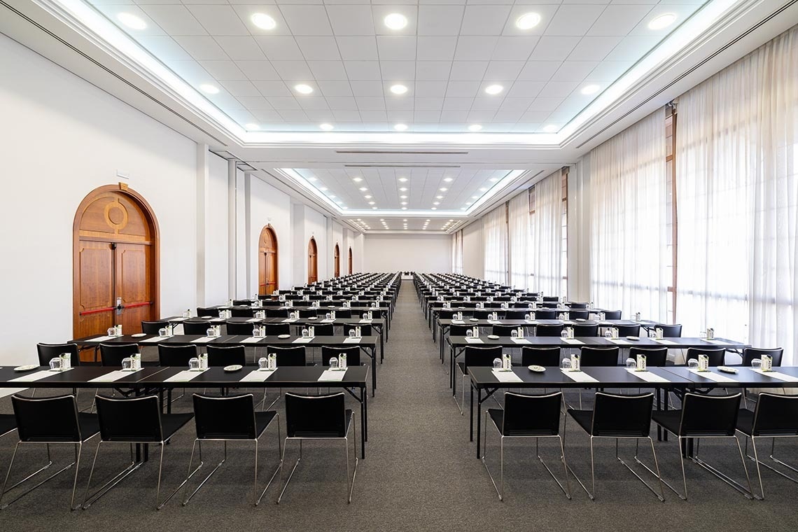 An empty, brightly lit conference room features rows of black chairs on a blue carpet, several side tables, and large arched windows offering views of a green landscape.