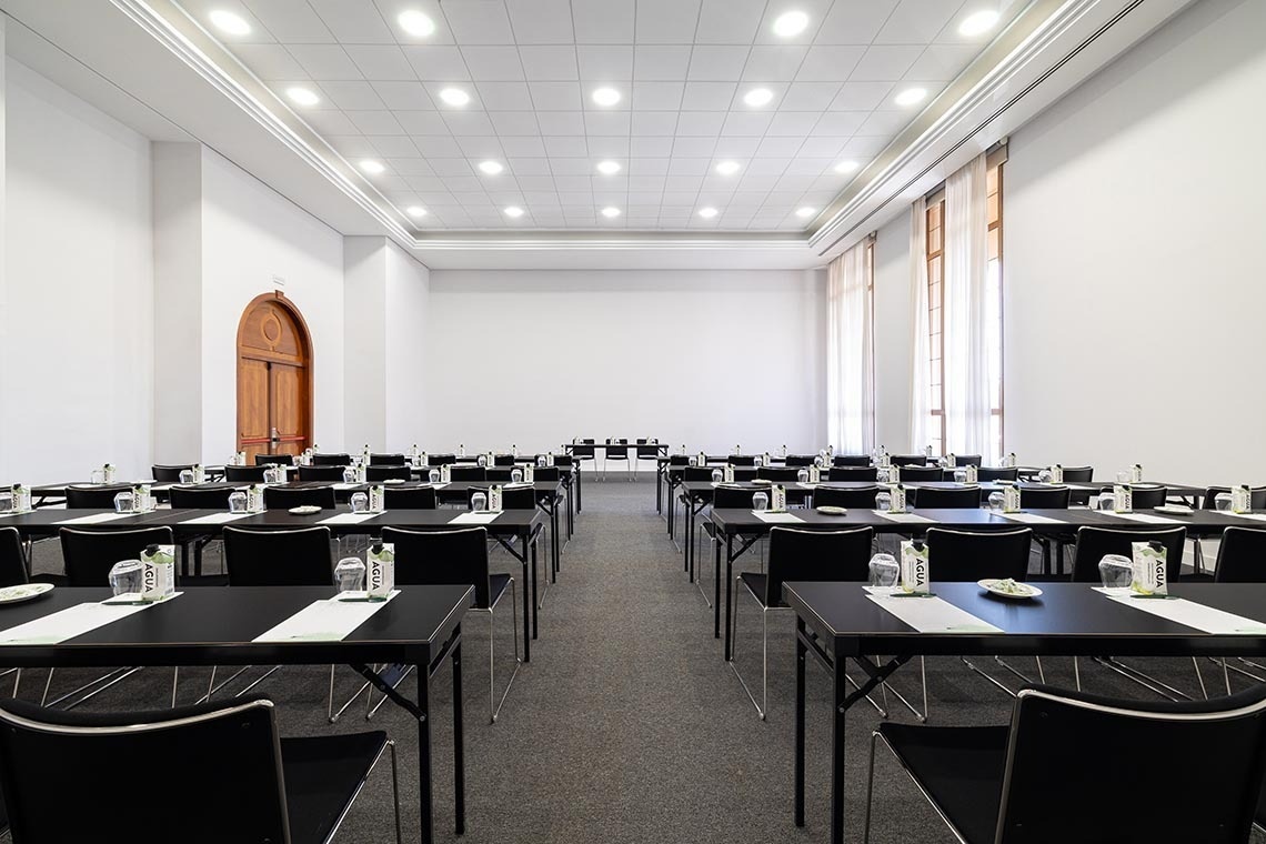 A spacious and well-lit conference room is meticulously set up with multiple rows of black tables and chairs, each place setting equipped with a bottle of water and a glass, and features an arched wooden door on one side and tall windows with sheer curtains on the other.