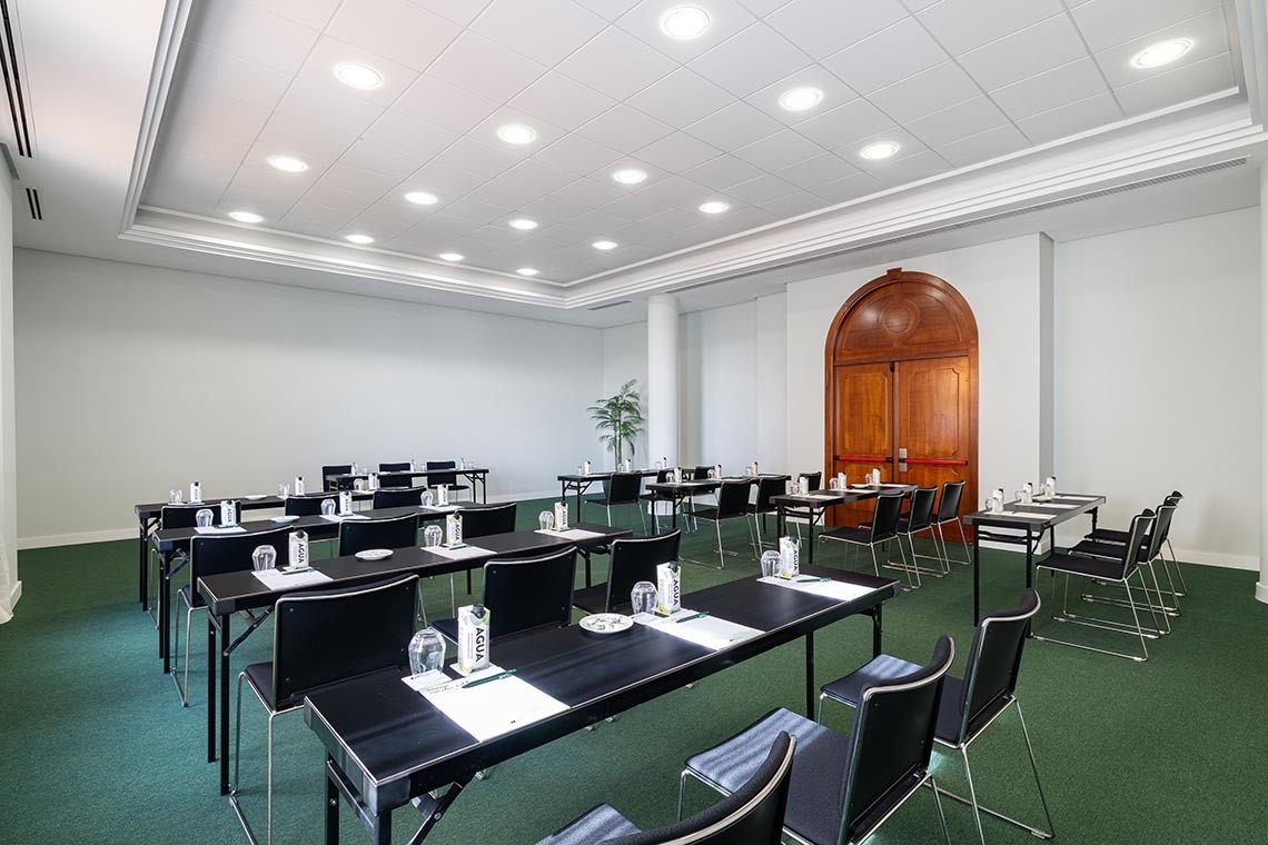 A well-lit conference room is arranged with rows of black tables and chairs, each set with water and paper, and features a prominent arched wooden door at one end.