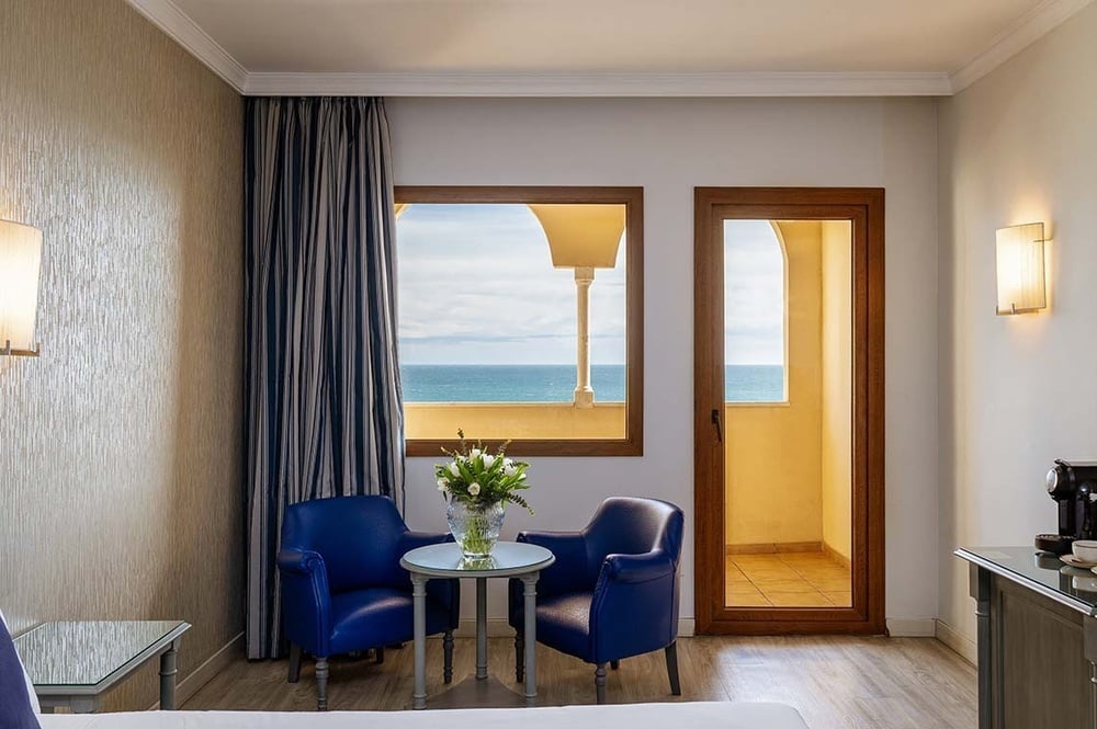 Das Bild zeigt ein helles Hotelzimmer mit Meerblick, ausgestattet mit zwei blauen Sesseln und einem kleinen Tisch mit frischen Blumen.